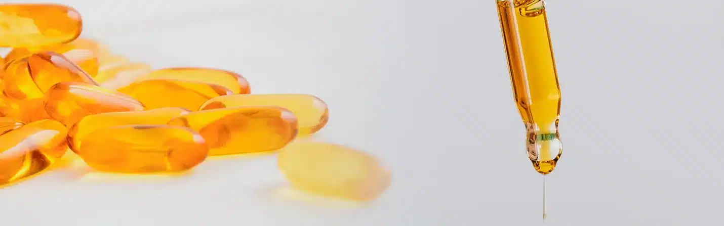 Amber softgel capsules on a white surface beside a glass dropper releasing a drop of oil